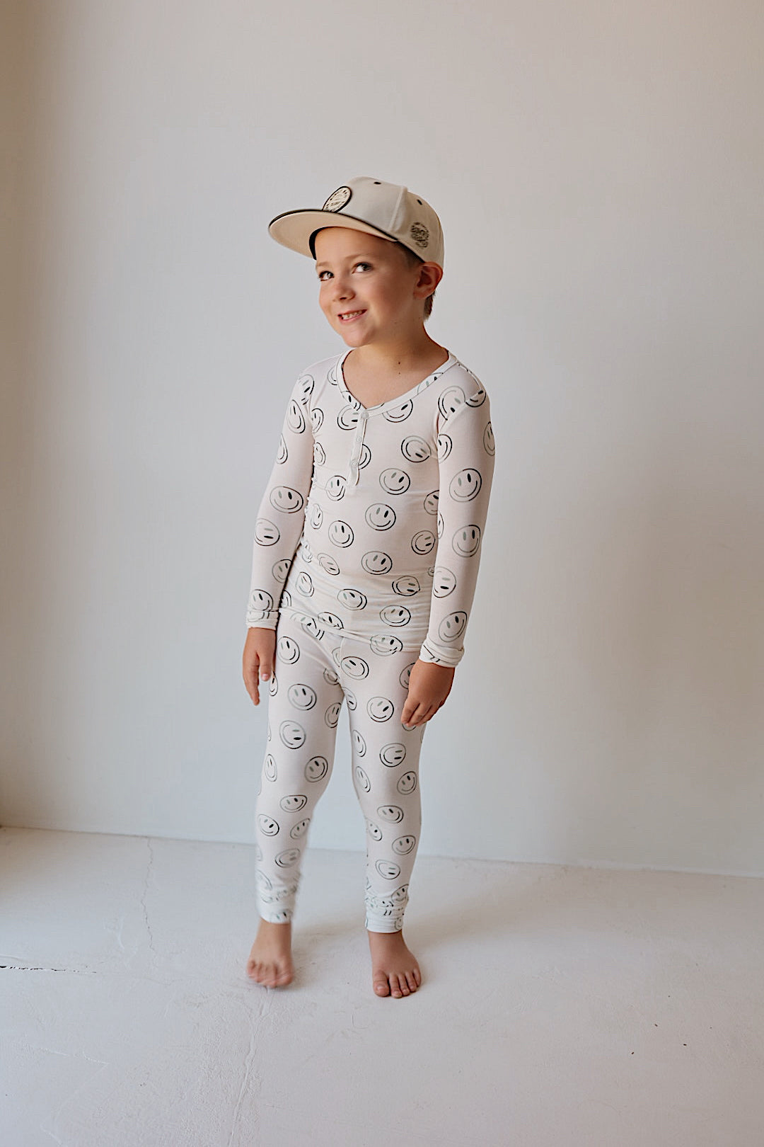 A young child stands barefoot against a plain wall, smiling and looking to the side, wearing forever french baby’s Bamboo Two Piece Pajamas | Green Smiles—lightweight, breathable sleepwear with a cheerful smiley face print.