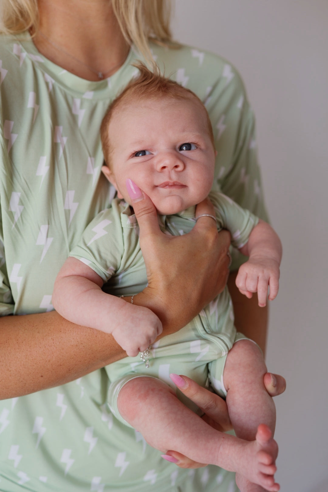 A baby wears the forever french baby Shortie Bamboo Zip | Flash, a light green romper with white lightning bolts, held by an adult in a matching shirt. The baby's curious look and the adult's pink nails highlight this hypoallergenic outfit.