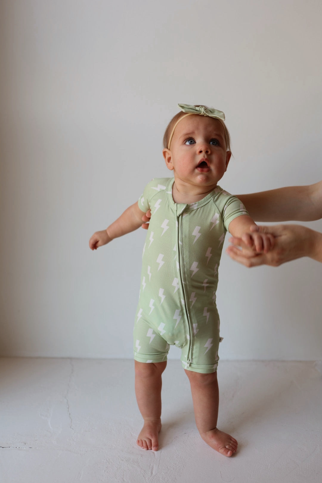 A toddler wears the forever french baby Shortie Bamboo Zip | Flash romper in light green with white lightning bolts and a matching headband, standing with support from an adult. The hypoallergenic material is gentle on sensitive skin. Plain, light-colored background.