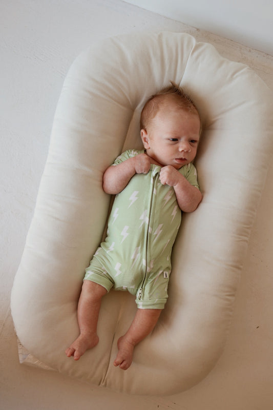 A light-skinned, red-haired baby lies on a soft cream cushion in the forever french baby Shortie Bamboo Zip | Flash romper—a light green bamboo outfit with white lightning bolts—looking to the side with hands near the zipper.