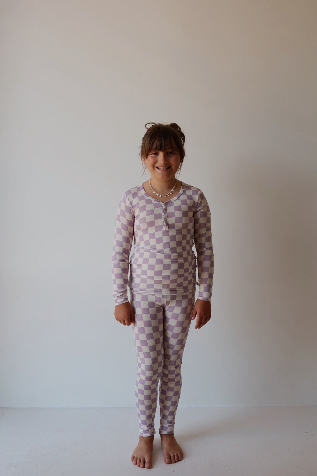 A young girl, barefoot and smiling against a light wall, wears the forever french baby Bamboo Two Piece Pajamas | Grape Soda—a breathable lavender and white kids' sleepwear set—paired with a beaded necklace.
