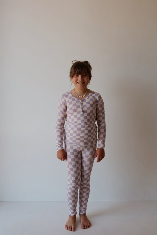 A young girl, barefoot and smiling against a light wall, wears the forever french baby Bamboo Two Piece Pajamas | Grape Soda—a breathable lavender and white kids' sleepwear set—paired with a beaded necklace.