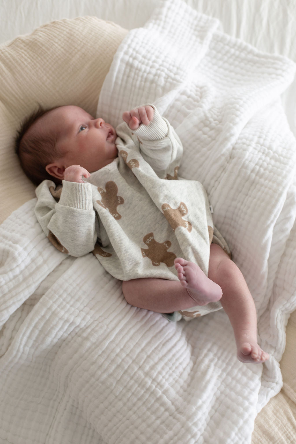 A newborn lies on a soft, white blanket wearing the forever french baby Sweatshirt Romper | Gingerbread 2.0, featuring gingerbread patterns. The baby looks to the side with hands near their face and feet slightly bent.