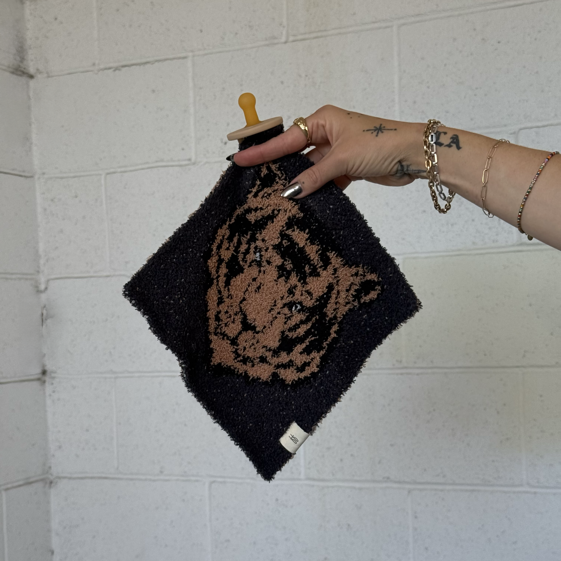 A tattooed hand holds the forever french baby Lovey | Tiger, a dark square microfiber blanket featuring a tan tiger face and attached black-and-tan pacifier, against a white brick wall.