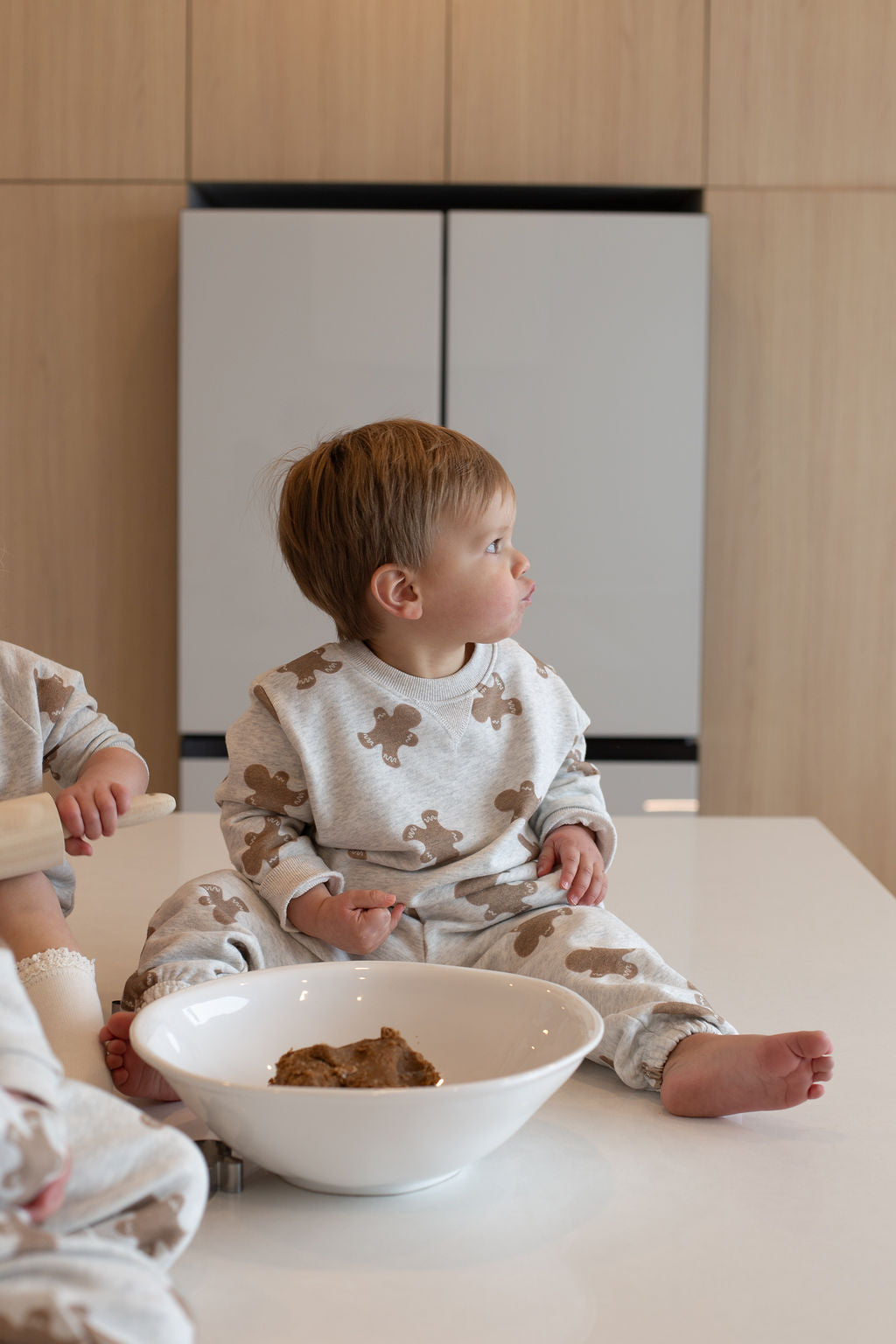 A toddler in the forever french baby Sweat Set | Gingerbread 2.0 sits barefoot on a kitchen counter near a white bowl, gazing to the side. Light wood cabinets are in the background, with another child partially visible, sharing everyday moments.