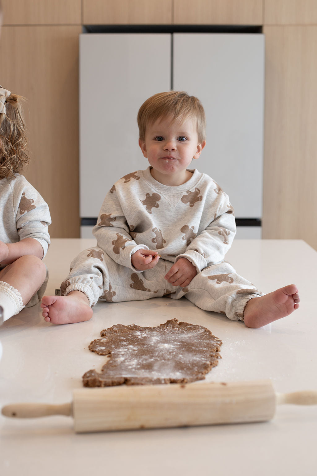 A young child in the forever french baby Sweat Set | Gingerbread 2.0 sits on a kitchen counter, smiling with dough and a rolling pin—perfectly dressed for everyday baking adventures.