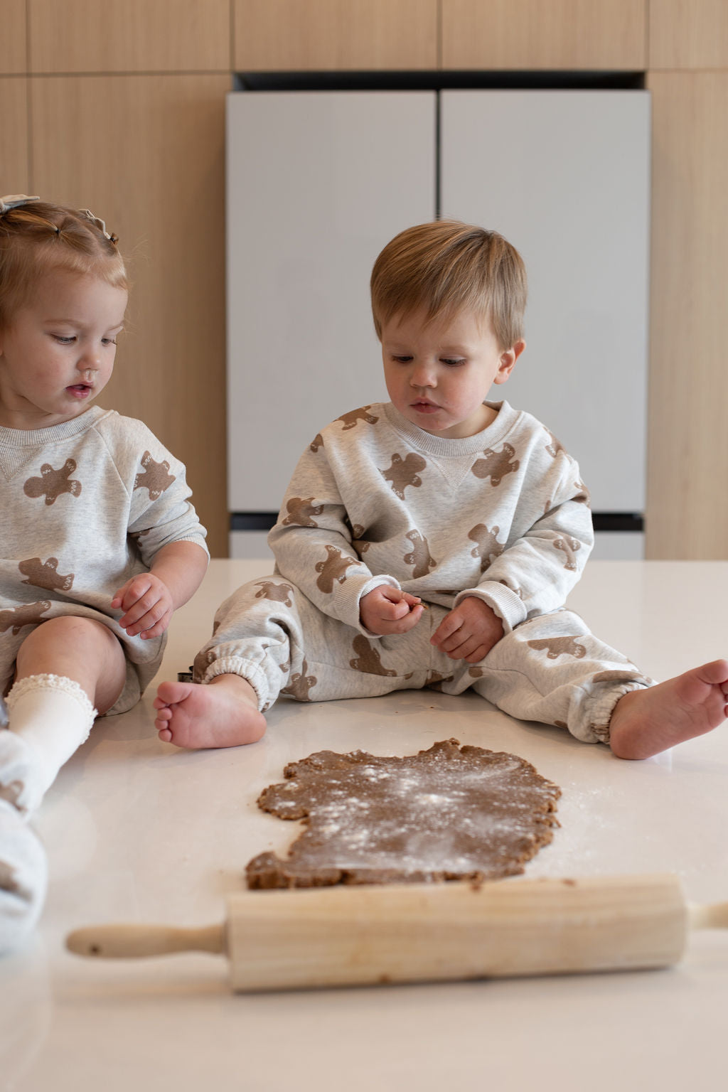 Two young children wear matching forever french baby Sweatshirt Romper | Gingerbread 2.0 sets as they sit on a bright kitchen counter, focused on gingerbread dough and a rolling pin in front of them.