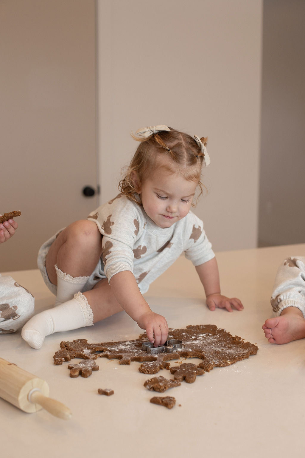 A young child wearing the forever french baby Sweatshirt Romper | Gingerbread 2.0 with brown spots and a hair bow sits on a counter, making cookie shapes with a cutter. Rolled-out dough, a rolling pin, and another child’s legs are nearby.