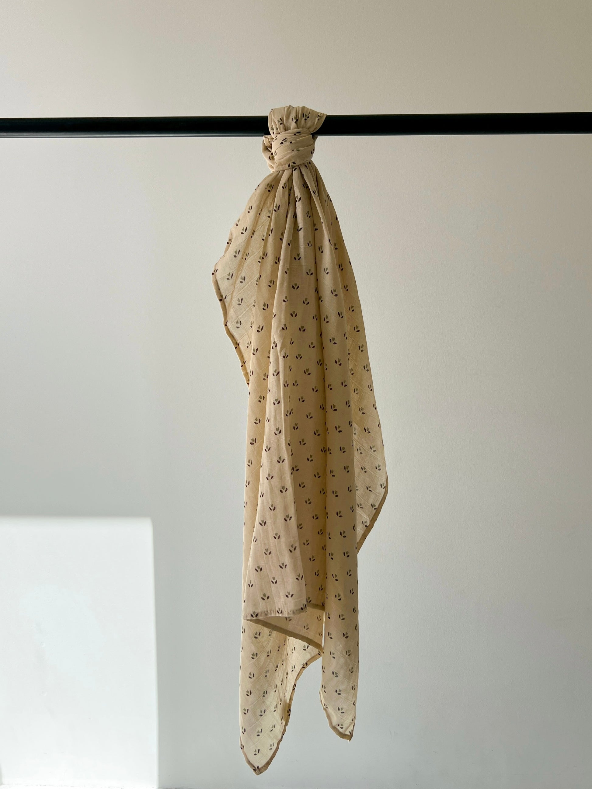 The Muslin Swaddle | Flora (Mis Print Peach Tone) by forever french baby, in a peach-toned muslin cotton with a delicate pattern, is draped over a black rod against a softly lit plain wall.