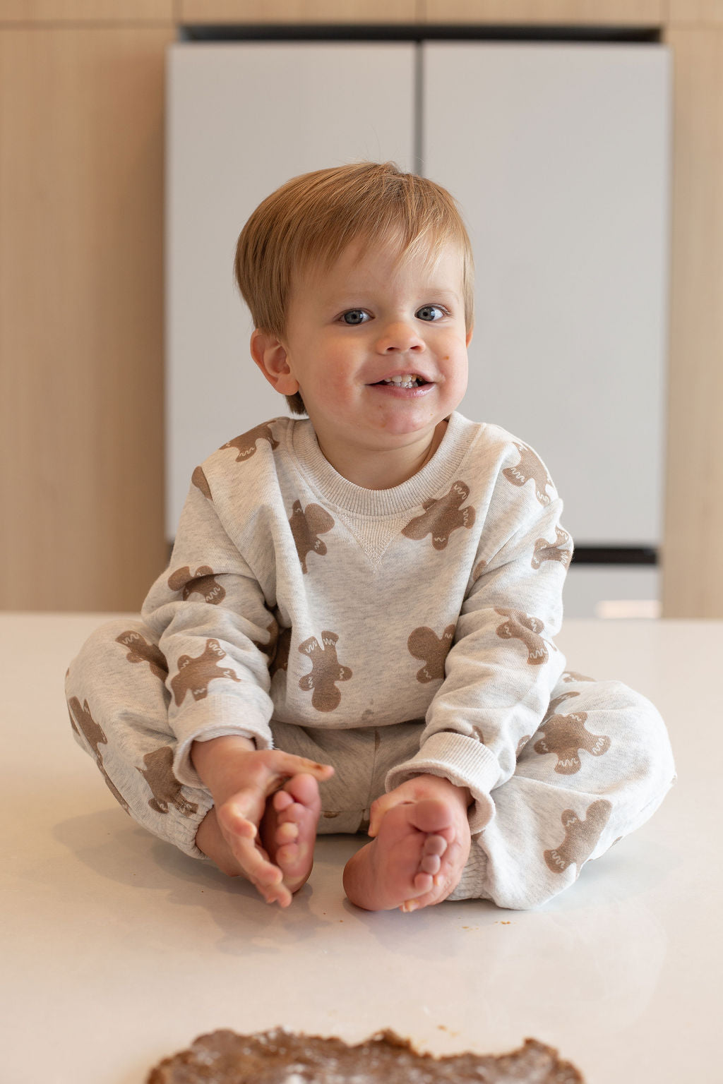 A smiling young child with light brown hair sits cross-legged on a white surface, wearing the forever french baby Sweat Set | Gingerbread 2.0 featuring brown teddy bear prints. Brown dough, echoing the gingerbread theme, is visible in the foreground.