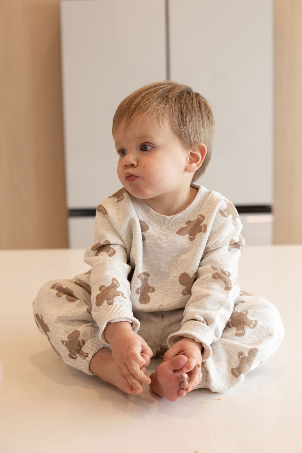 A light-haired child sits cross-legged on a white surface, wearing the forever french baby Sweat Set | Gingerbread 2.0, featuring playful prints and a curious expression—perfect for everyday adventures.