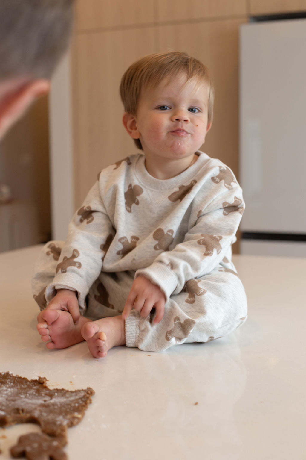 A toddler wears the forever french baby Sweat Set | Gingerbread 2.0, sitting barefoot on a kitchen counter with cookie dough nearby, gazing up playfully—capturing the everyday magic in cozy gingerbread-print style.