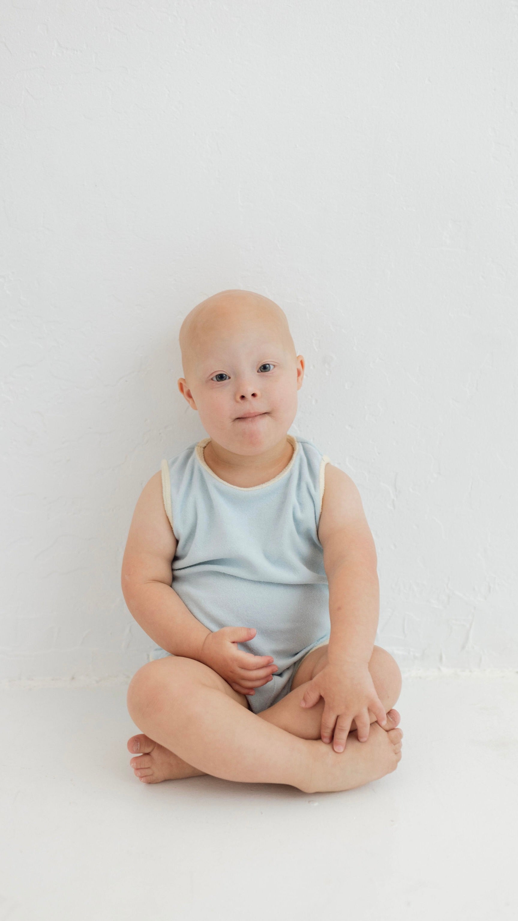 A young toddler with short hair sits on the floor against a plain white background, wearing the Blue Terry Tank Set by forever french baby, with a gentle expression and hands resting on crossed legs.