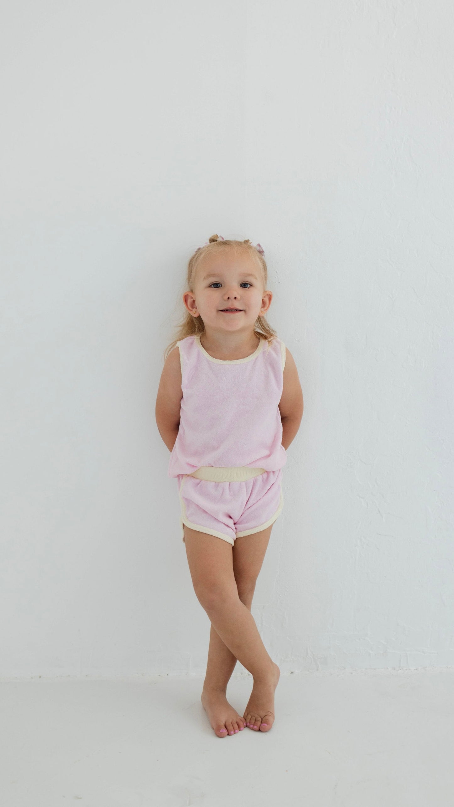 A young child with light hair, dressed in a Forever French Baby pastel purple Terry Tank Set, stands against a white wall with crossed legs and hands behind their back. They are smiling slightly and have small bows in their hair.