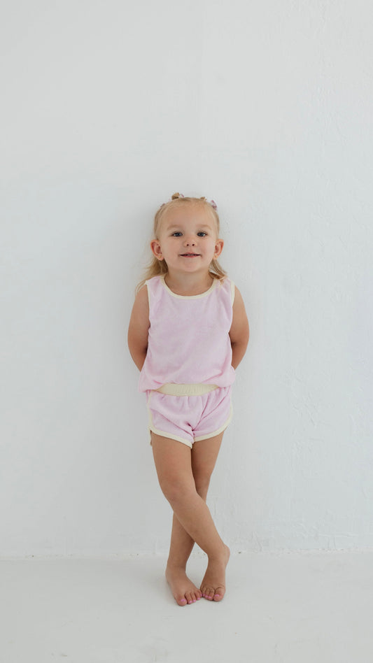 A young child with light hair, dressed in a Forever French Baby pastel purple Terry Tank Set, stands against a white wall with crossed legs and hands behind their back. They are smiling slightly and have small bows in their hair.