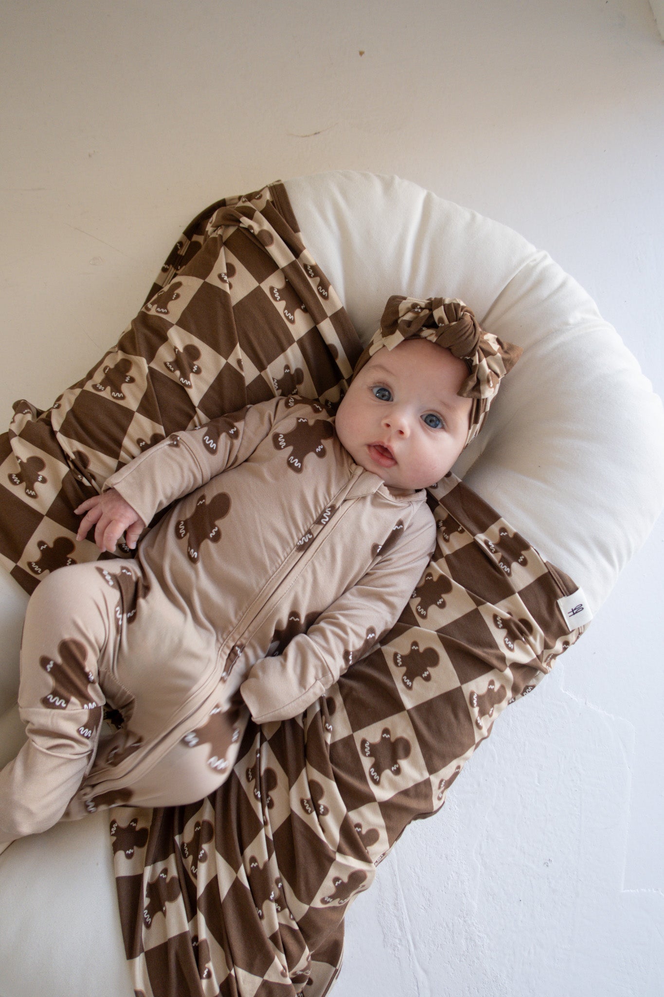 A baby clad in a brown outfit with bear prints is resting on a cushioned blanket that showcases a brown and white diamond pattern. The infant, with wide eyes peering upward, is adorned with an adorable bow from the Bamboo Head Wrap in Gingerbread Checkerboard by forever french baby.