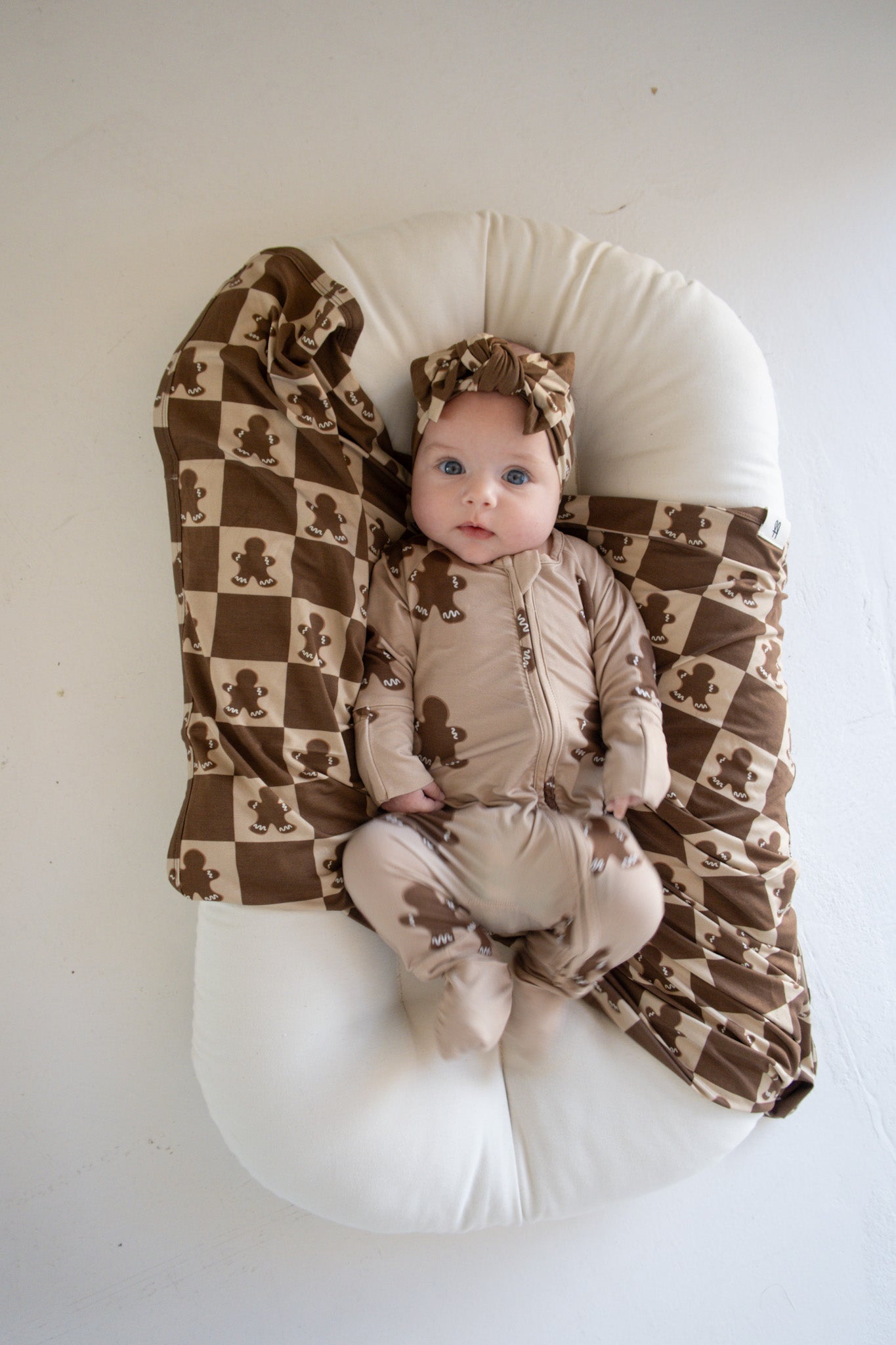 A baby lies on a white cushion, wearing a brown and beige outfit adorned with a teddy bear pattern. The matching blanket and adjustable Bamboo Head Wrap in Gingerbread Checkerboard, both from forever french baby, featuring alternating teddy bear designs, complete the look. The baby gazes up with a calm expression.