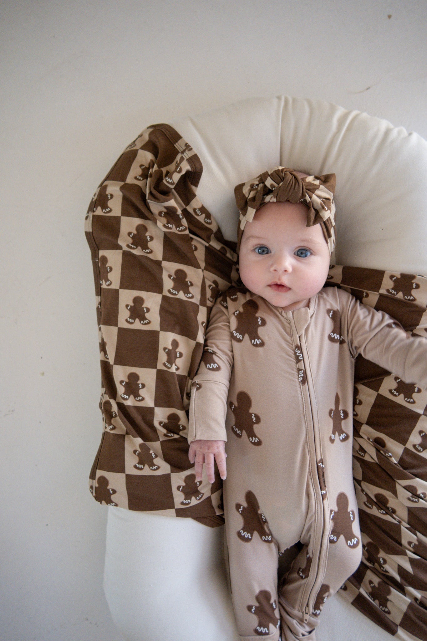 A baby wearing a brown, patterned onesie and matching headband lies on a cream cushion. The hypo-allergenic Bamboo Swaddle | Gingerbread Checkerboard by forever french baby features a checkerboard pattern with teddy bears. The baby has bright blue eyes and gazes at the camera.