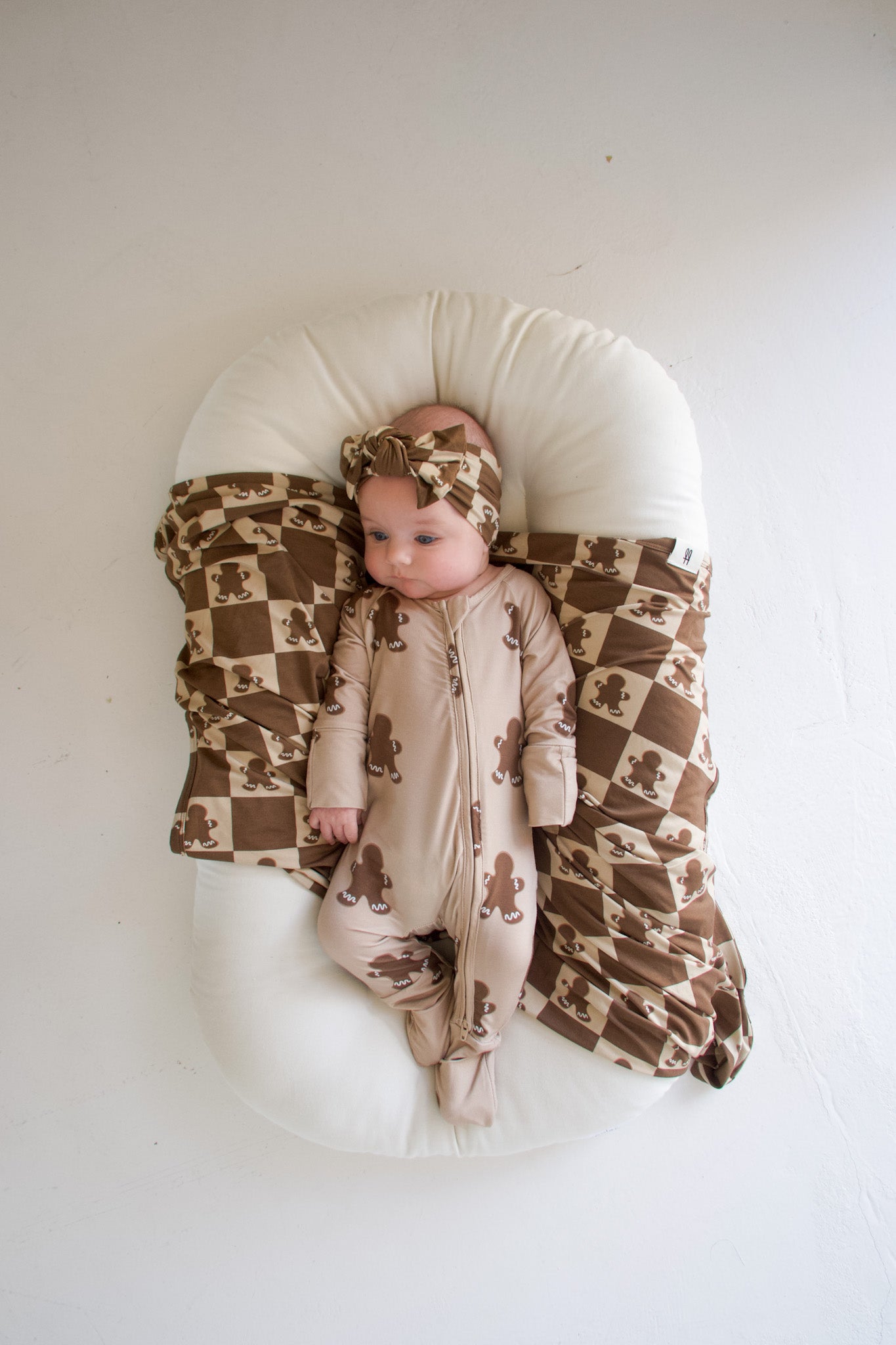 A baby lies comfortably on a white cushion, dressed in a brown outfit adorned with teddy bear patterns and complemented by a matching headband. The hypo-allergenic cushion is partially draped with the forever french baby's Bamboo Swaddle in the Gingerbread Checkerboard design. The baby looks to the side, nestled cozily in breathable fabric.