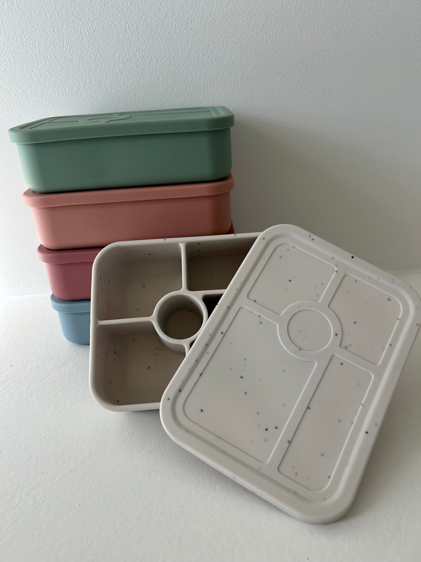 A stack of colorful Bento Boxes by forever french baby, made from BPA-free silicone, arranged against a white background. The top box is green, followed by pink, blue, and gray boxes. One gray Bento Box is open, revealing its divided compartments inside.