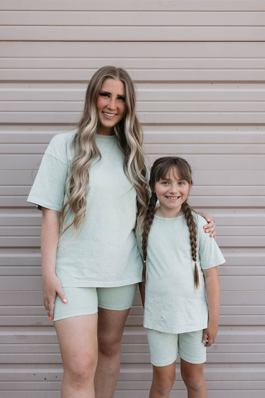 A woman with long blonde hair and a young girl with brown hair in pigtails stand side by side, smiling at the camera in matching light green outfits. The girl is wearing an adorable Children's Short Set | Vintage Washed Spearmint from forever french baby, while the woman has her arm around her shoulders. They pose in front of a beige, horizontal-panel wall.