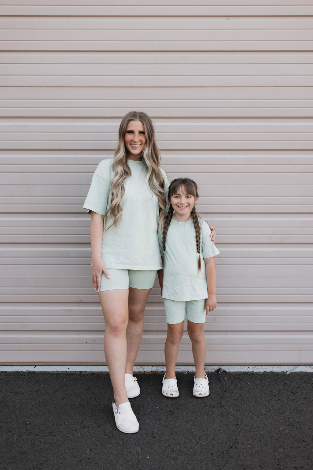 A woman and a young girl stand in front of a beige garage door, both matching in their Women's Short Sets from forever french baby in Vintage Washed Spearmint. The woman, with long wavy hair, has her arm around the girl, who wears her hair in two braids. Both are smiling and have completed their outfits with white shoes.