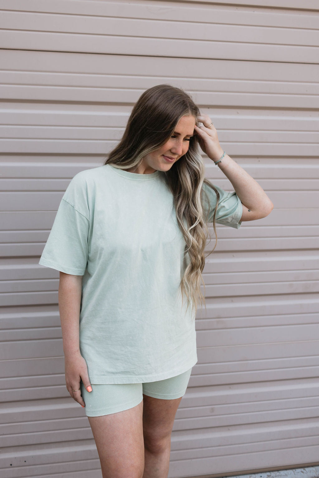 A person with long, wavy hair stands in front of a beige, horizontal-paneled wall. They are wearing a loose-fitting Women's Short Set in Vintage Washed Spearmint from forever french baby. Their posture is relaxed, with one hand touching their hair.