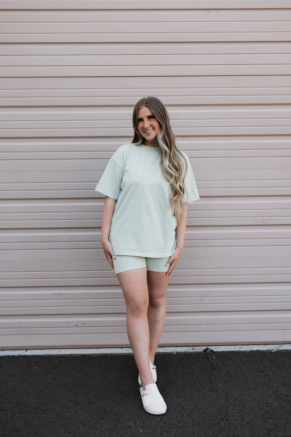 A woman stands in front of a beige garage door, smiling with her hands relaxed by her sides. She is wearing a Vintage Washed Spearmint Women's Short Set from forever french baby—an oversized mint green t-shirt paired with matching biker shorts, white slip-on shoes, and has long, wavy blonde hair.