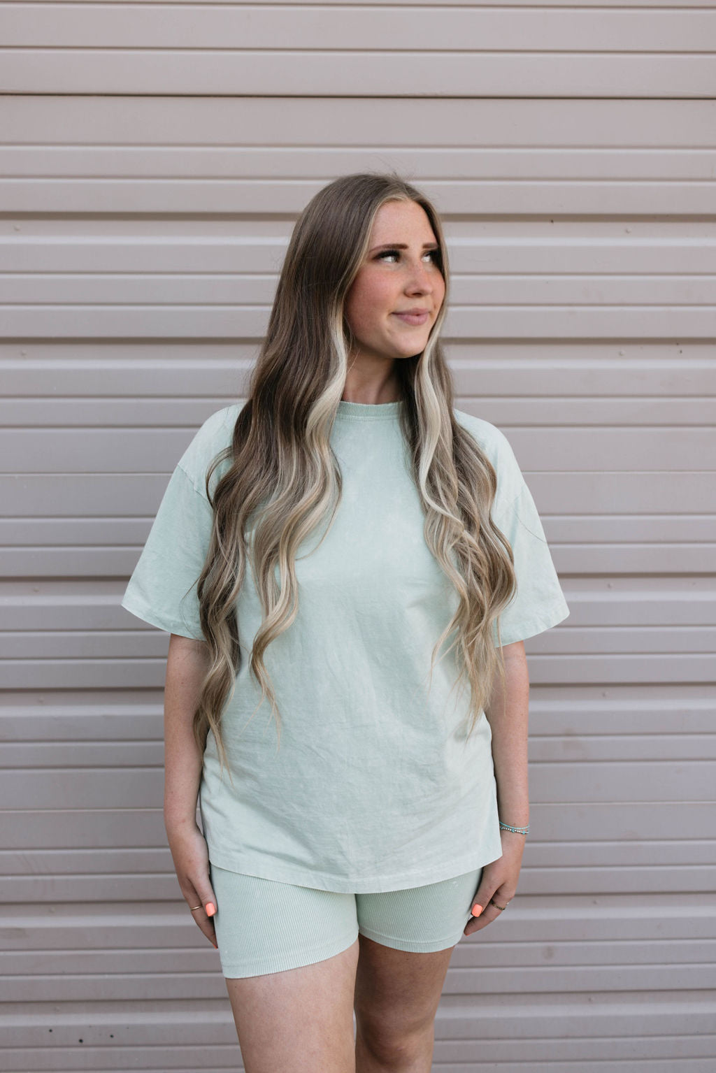 A person with long, wavy hair stands against a light gray, ridged backdrop. They are wearing the Women's Short Set in Vintage Washed Spearmint by forever french baby, looking slightly to their right with a relaxed expression.