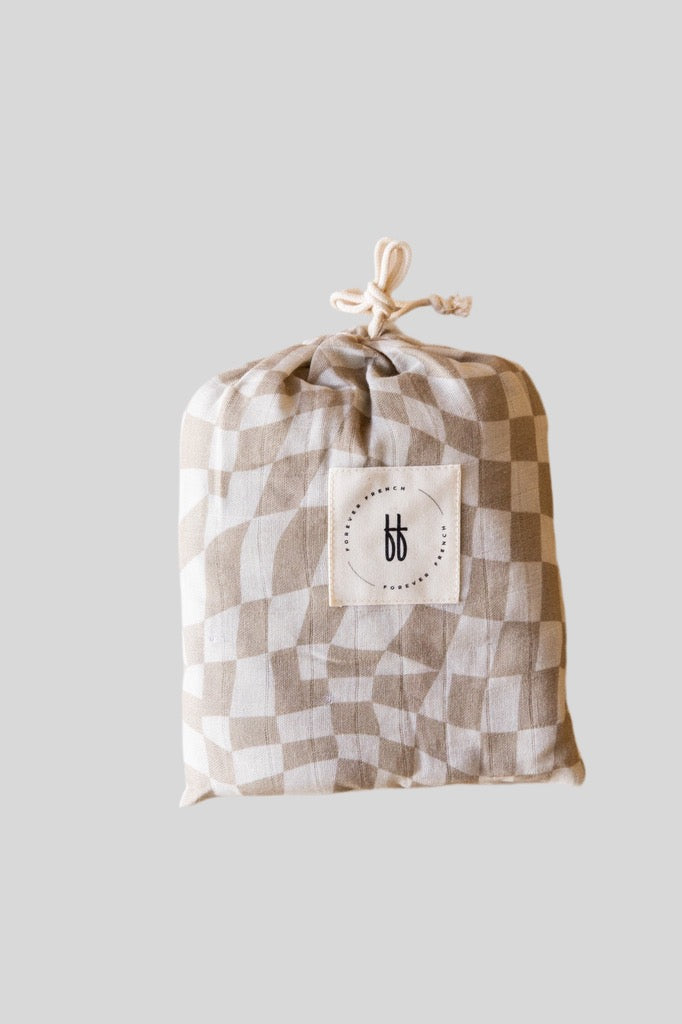 A muslin cotton fabric pouch in beige and white wavy checkerboard pattern with a drawstring closure at the top. The pouch from forever french baby features a white square label on the front, showcasing a minimalist black logo with stylized lowercase "bb" letters.