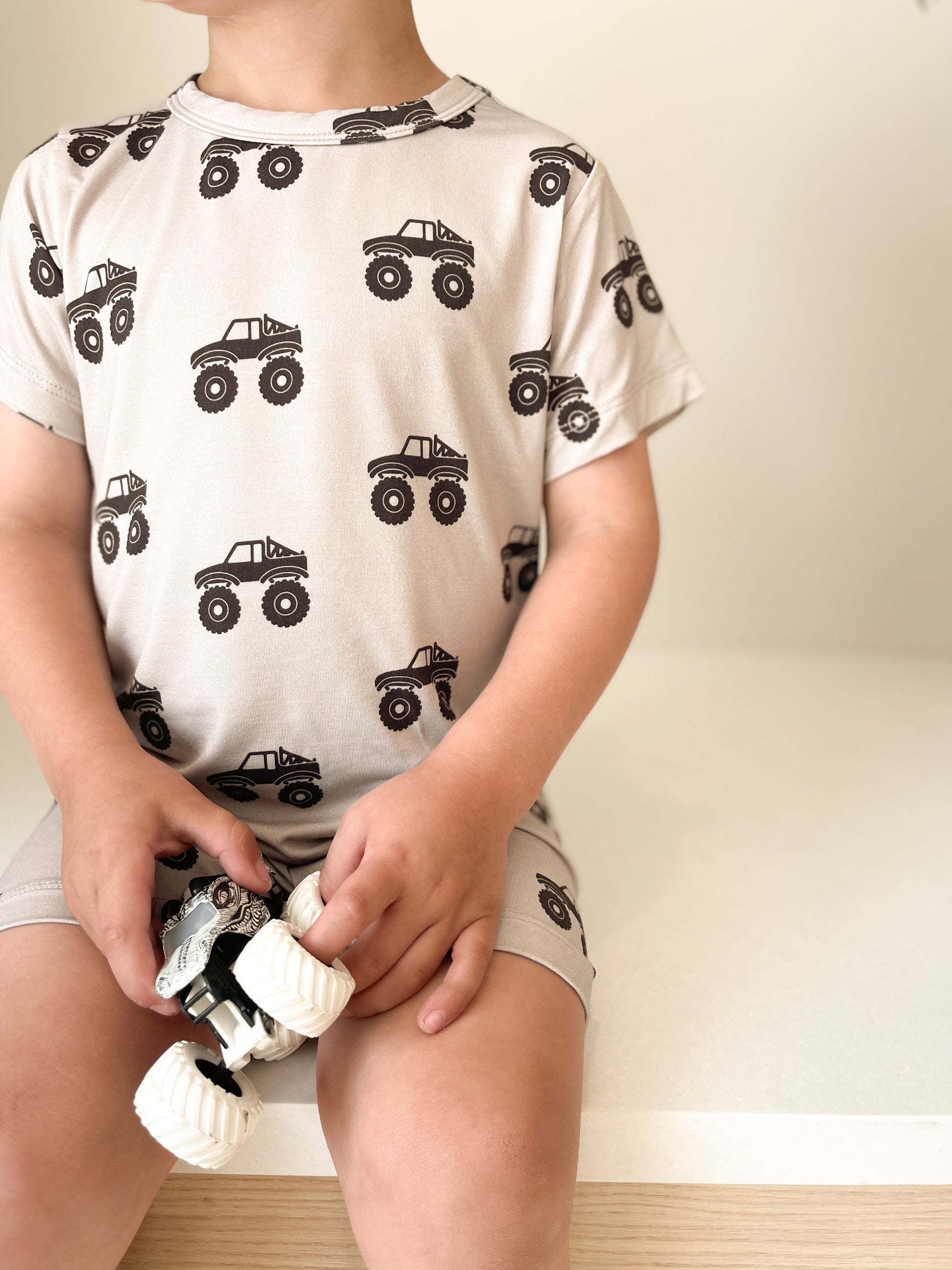 A young child, face not visible, sits on a light surface wearing the forever french baby Bamboo Short Set | Monster Truck, holding a small white toy vehicle. The set features breathable fabric with black monster truck prints.