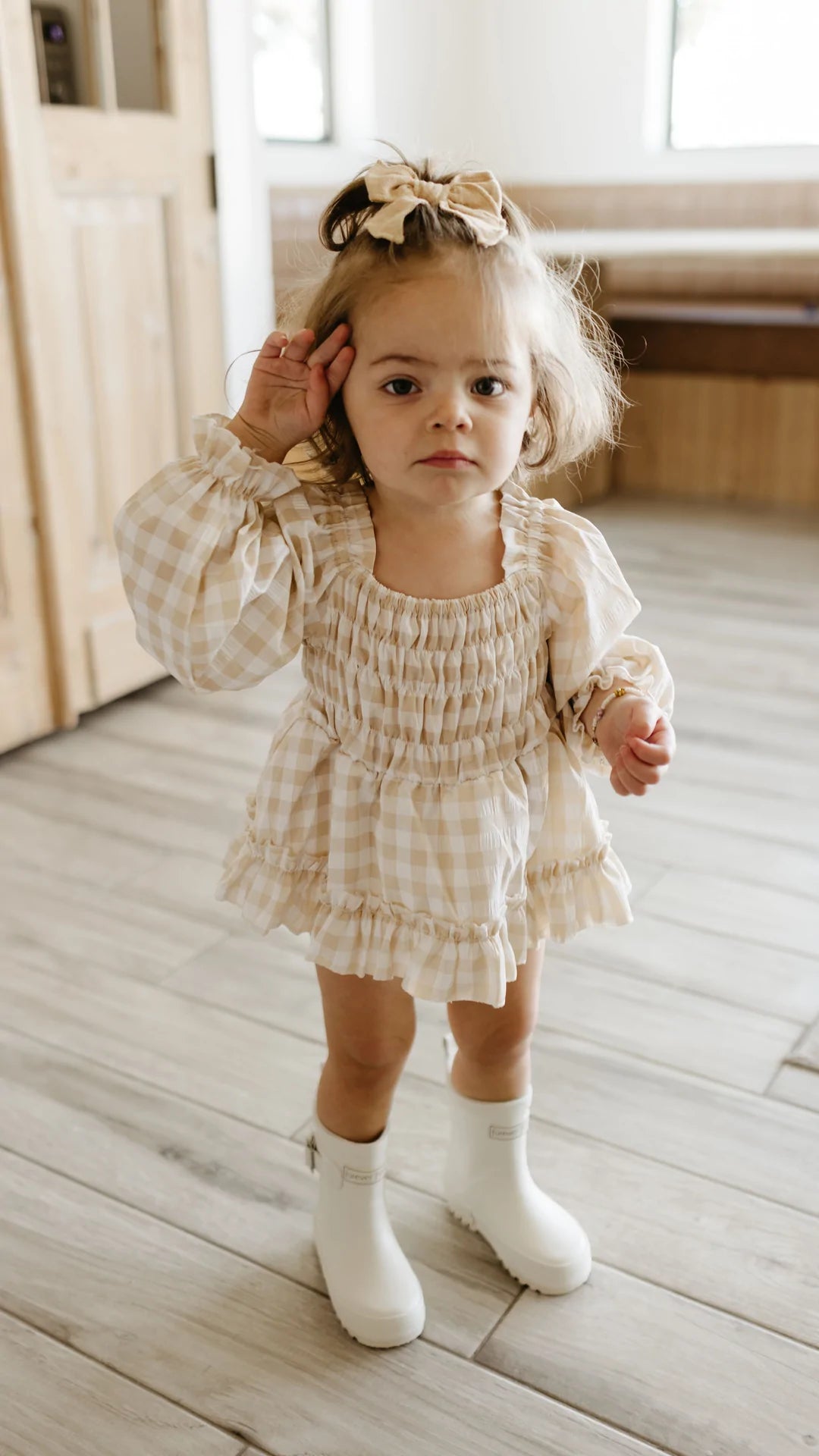 A toddler wearing a Ruffle Romper in beige gingham from forever french baby and white boots stands indoors. They have a beige bow in their hair and are making a curious gesture with one hand near their ear. The room’s wooden flooring and light-colored walls perfectly complement the charming outfit.