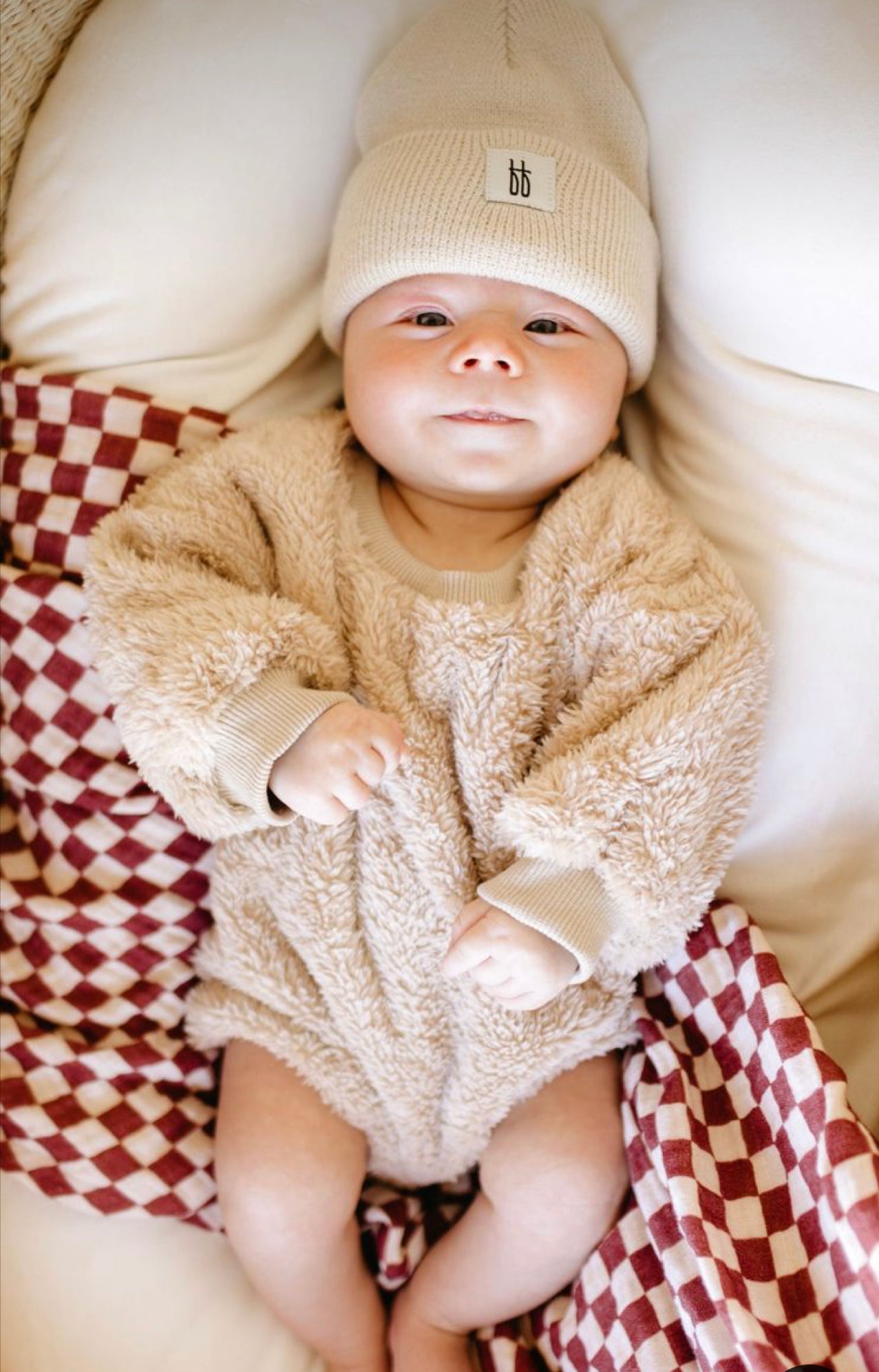 A baby in a cozy "Teddy Bear" long sleeve romper by forever french baby and a matching beanie lies on a white pillow, surrounded by a red and white checkered blanket. The baby has a content expression and clenched fists, looking up at the camera.