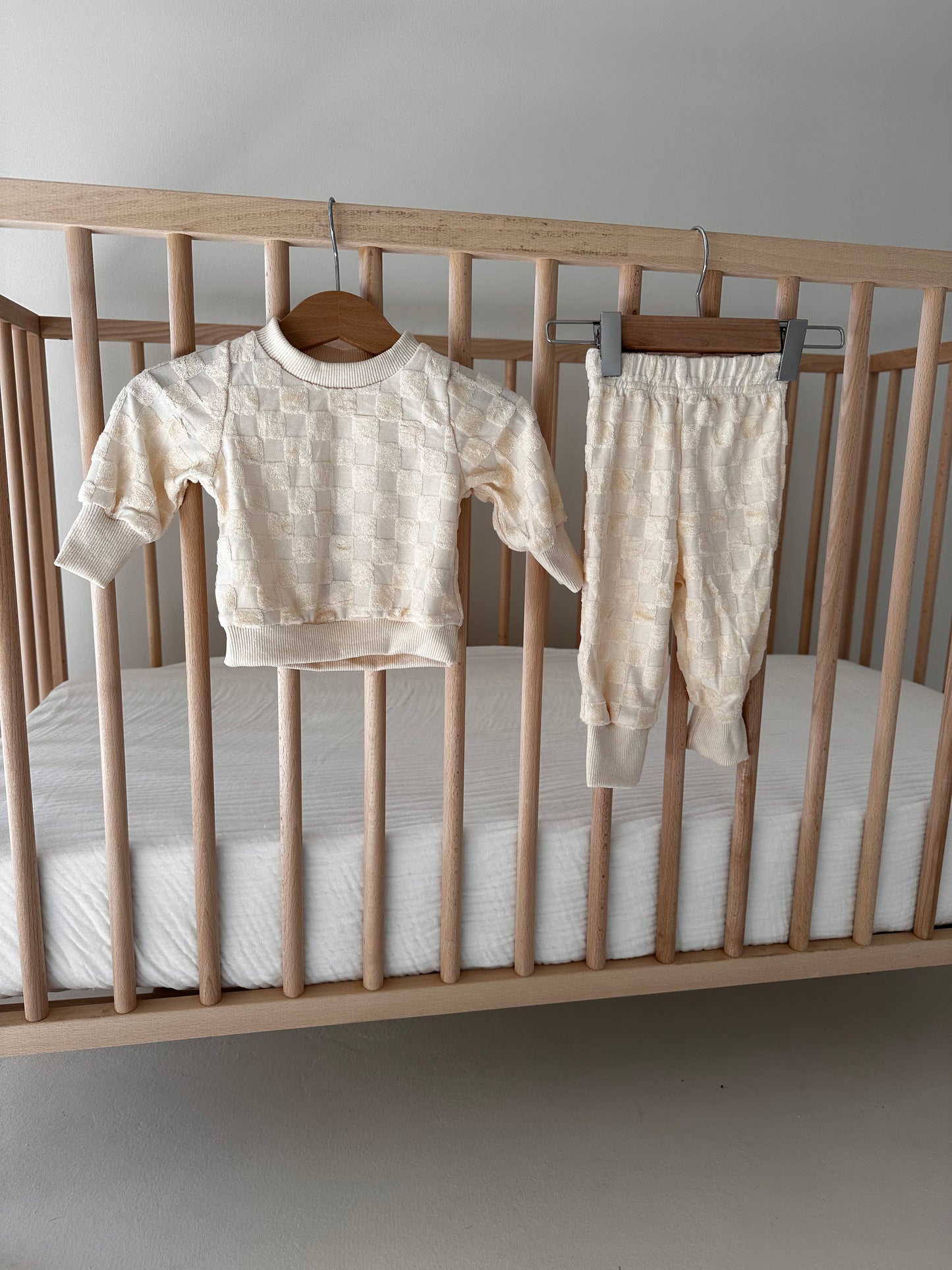 A light wooden baby crib holds a white mattress. Hung on the crib is a cream-colored Long Sleeve Set | Cream Terry Checkerboard from forever french baby, featuring a subtle textured pattern. The outfit is displayed neatly, highlighting its soft, cozy material.