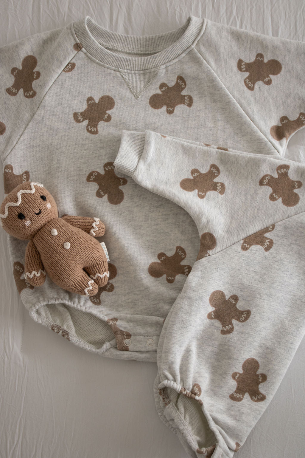 The forever french baby Sweatshirt Romper | Gingerbread 2.0 features a cozy, light gray design with brown gingerbread prints, paired with a small knitted gingerbread doll—ideal for your newborn collection and displayed on a white fabric background.