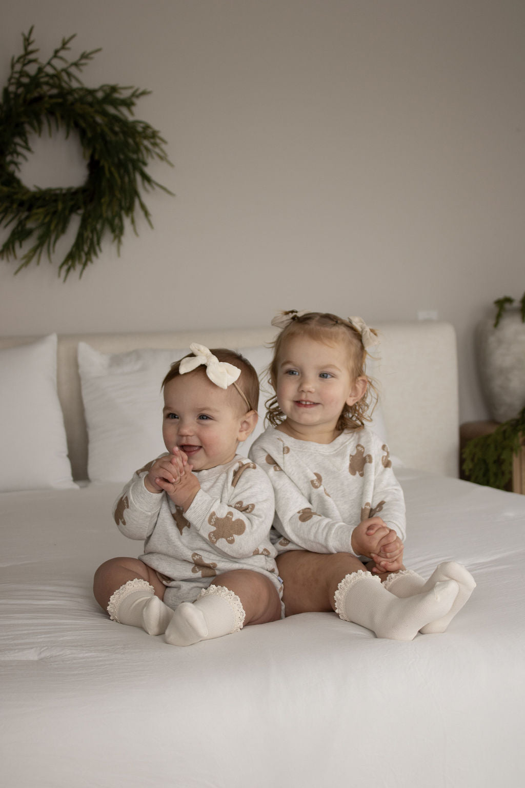Two young children with light hair wear matching Sweatshirt Romper | Gingerbread 2.0 by forever french baby and white socks on a white bed. One has a bow headband. A green wreath decorates the wall behind them.