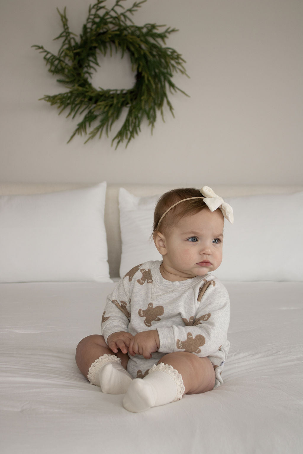 A baby in the forever french baby Sweatshirt Romper | Gingerbread 2.0 with gingerbread prints, cream socks, and a white bow headband sits on a bed with white linens; a green wreath hangs on the wall behind.