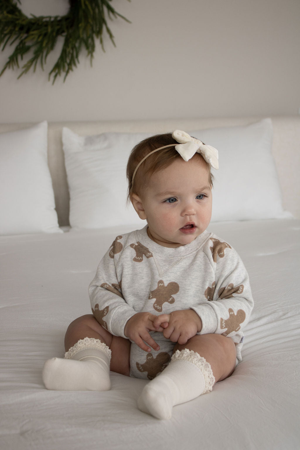 A light-haired baby in a cream bow headband sits on a white bed, wearing the forever french baby Sweatshirt Romper | Gingerbread 2.0 with cream socks. Soft white pillows and a green wreath decorate the background.