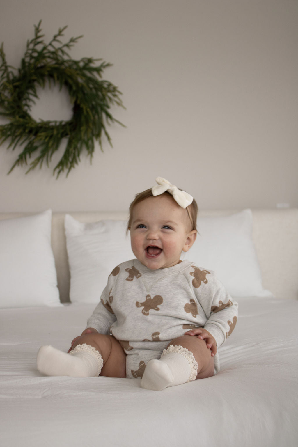 A smiling baby in the forever french baby Sweatshirt Romper | Gingerbread 2.0, with brown gingerbread figures, white socks, and a white bow headband sits on a bed with white pillows while a green wreath hangs on the wall behind.