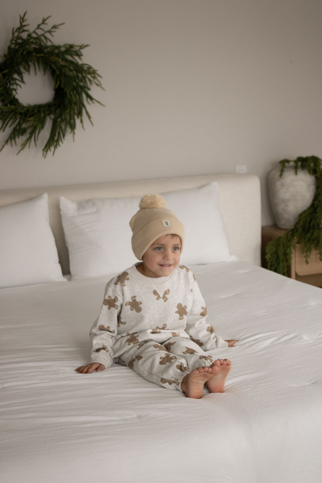 A young child in the forever french baby Sweat Set | Gingerbread 2.0 and a beige beanie smiles on a white bed in a softly lit, cozy bedroom with greenery decorating the wall and furniture.