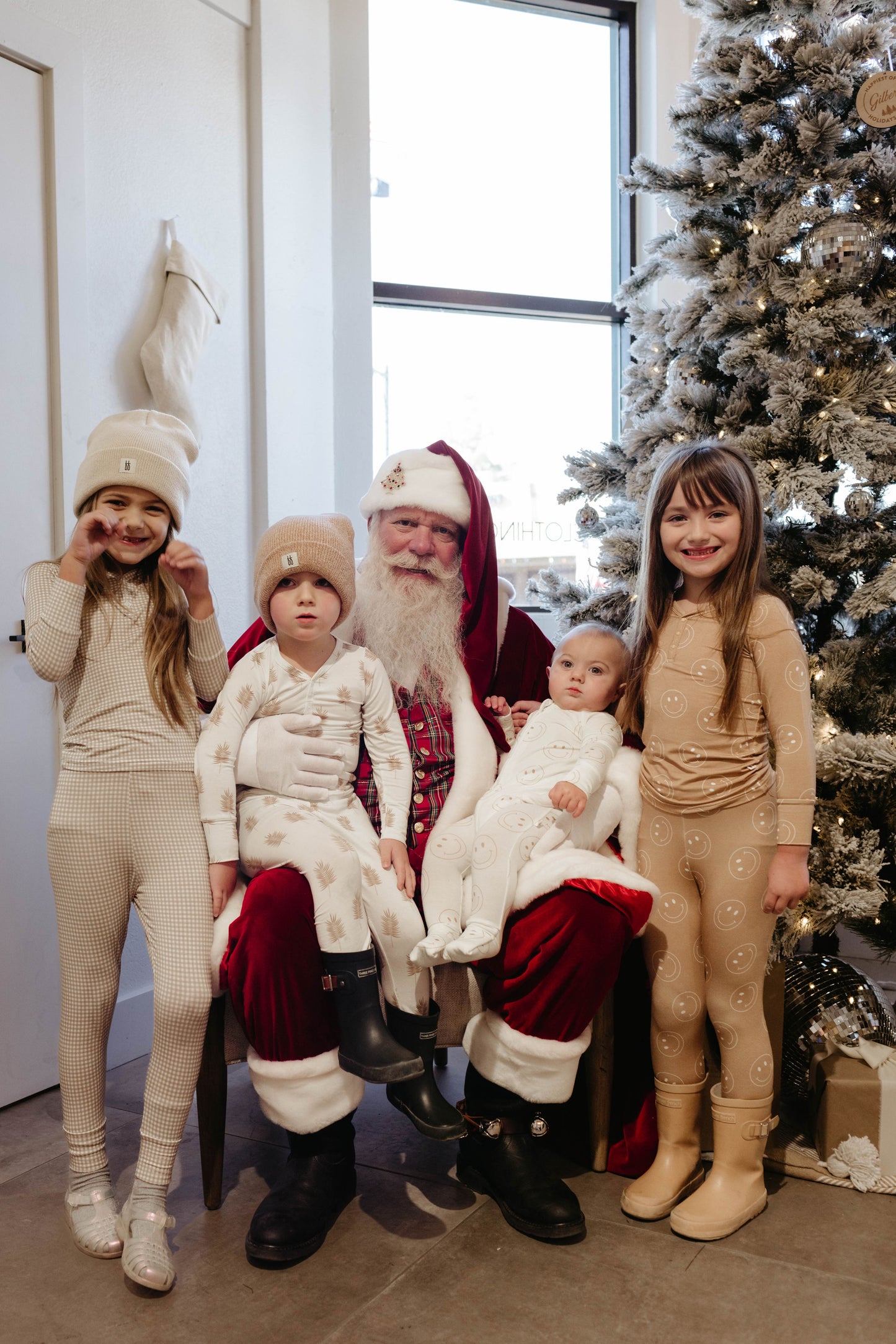 A heartwarming scene captured by Amanda Riley Photos, titled "Santa Photos | Forever French Baby x Amanda Riley Photos," features a man dressed as Santa Claus sitting near a decorated Christmas tree with four children. The children, clad in light-colored pajamas and winter hats from Forever French Baby, smile and pose around Santa in this festive setting.