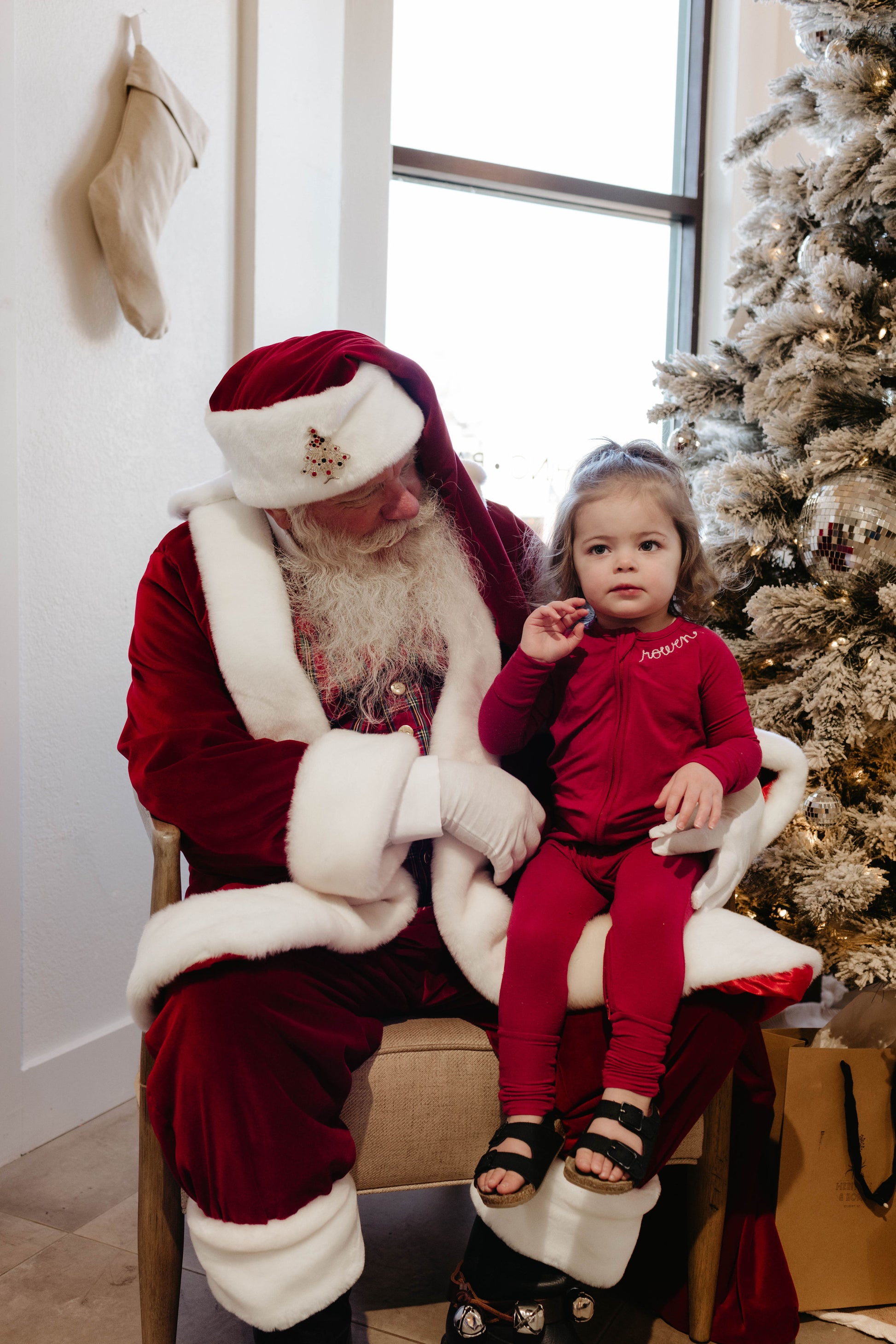 A child in red pajamas sits on Santa Claus' lap in a cozy room decorated for Christmas, captured by Amanda Riley Photos. This joyful moment near a frosted Christmas tree adorned with decorations and a white stocking hanging on the wall in the background is brought to life by the collaboration of Santa Photos | Forever French Baby x Amanda Riley Photos.