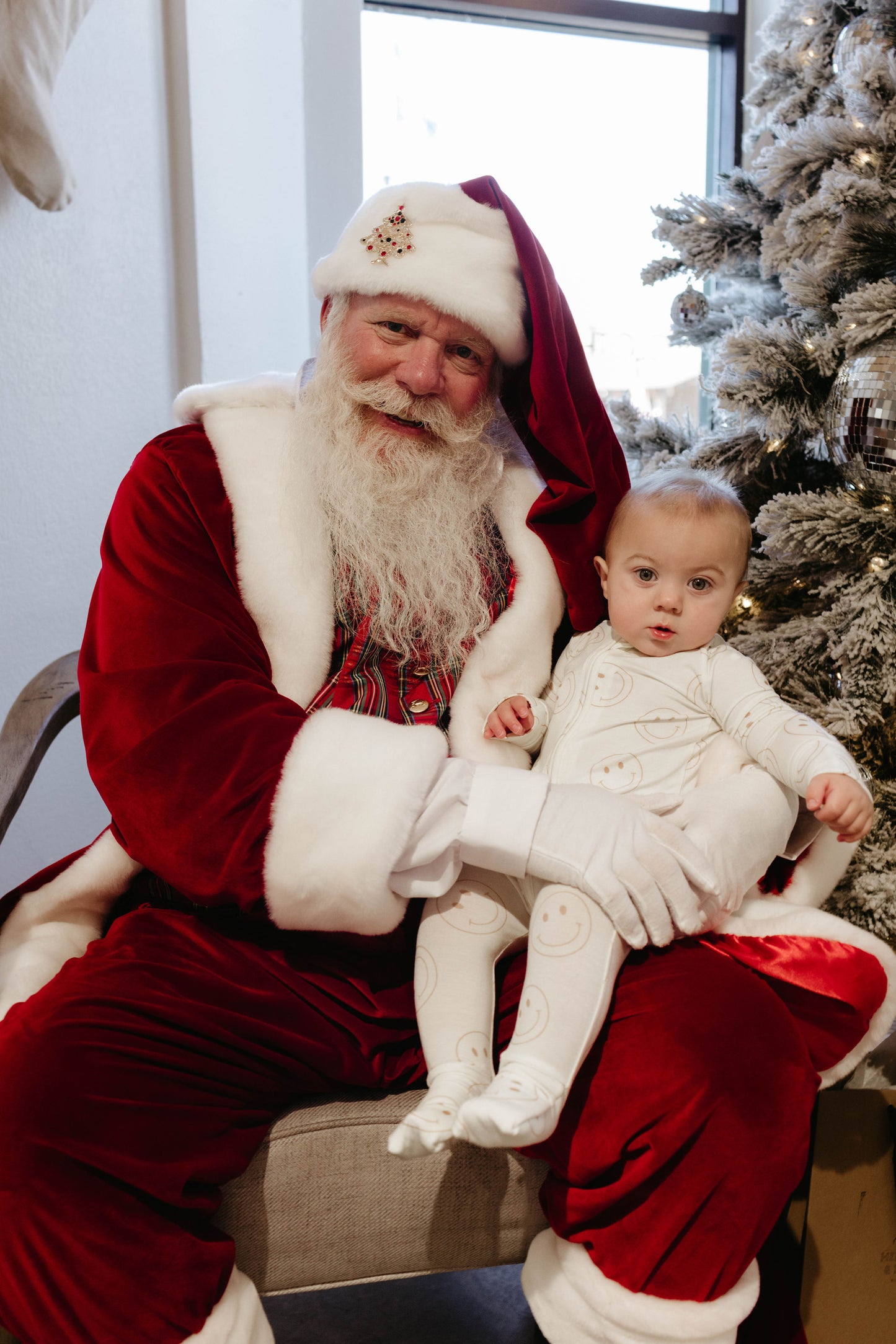 A man dressed as Santa Claus, adorned with a white beard, red suit, and hat, holds a baby clad in white pajamas decorated with smiley faces. Seated on an armchair next to a festively decorated Christmas tree, the baby gazes curiously at the camera. This scene perfectly captures the enchanting moments of Amanda Riley Photos' Santa Photos | Forever French Baby x Amanda Riley Photos collection.