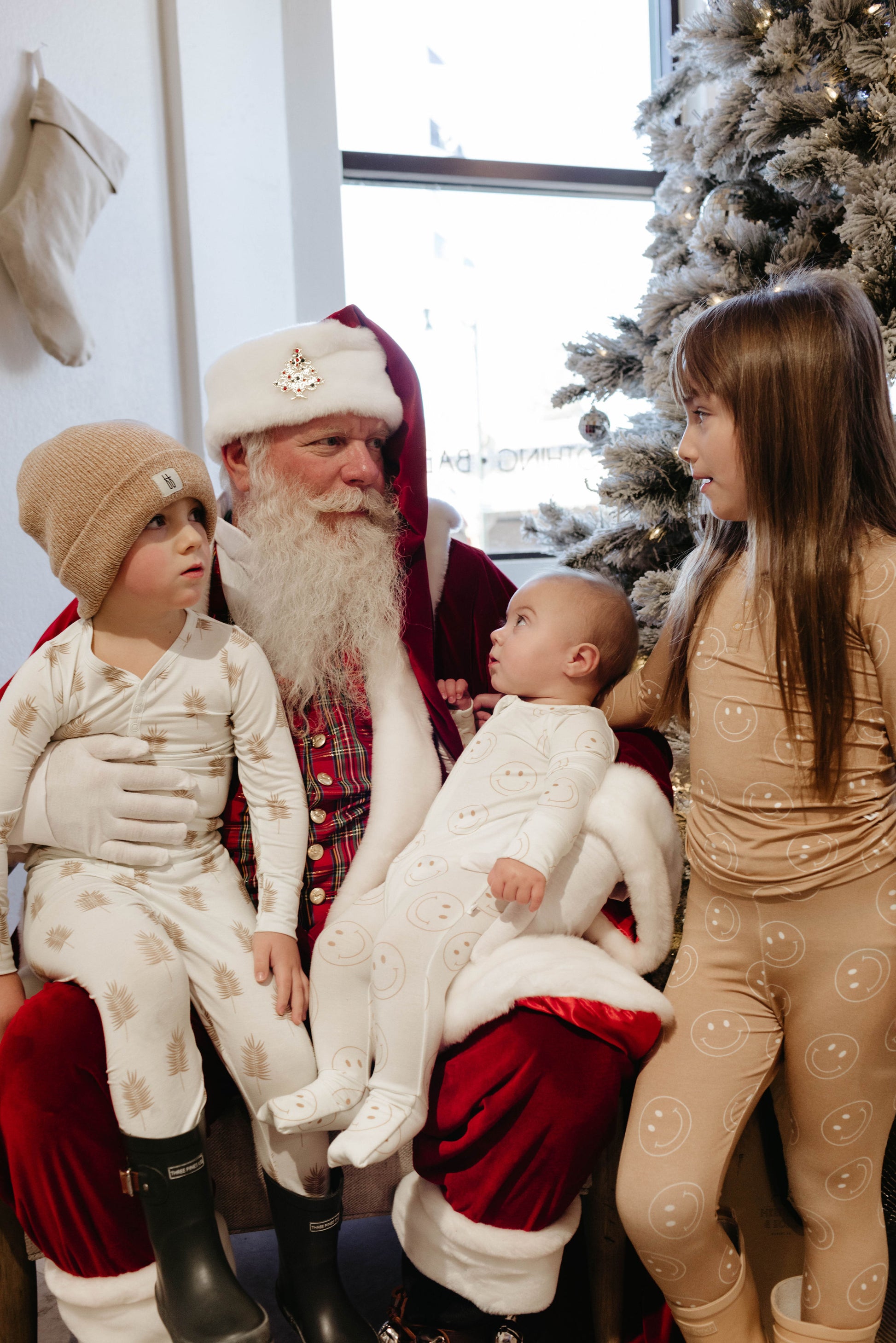 A person dressed as Santa Claus sits on a chair holding a baby in white pajamas, with another child in white pajamas and a tan beanie on the left, and a child in tan pajamas standing on the right near a decorated Christmas tree. This charming scene is perfectly captured by Amanda Riley Photos as part of the Santa Photos collection from Forever French Baby x Amanda Riley Photos.