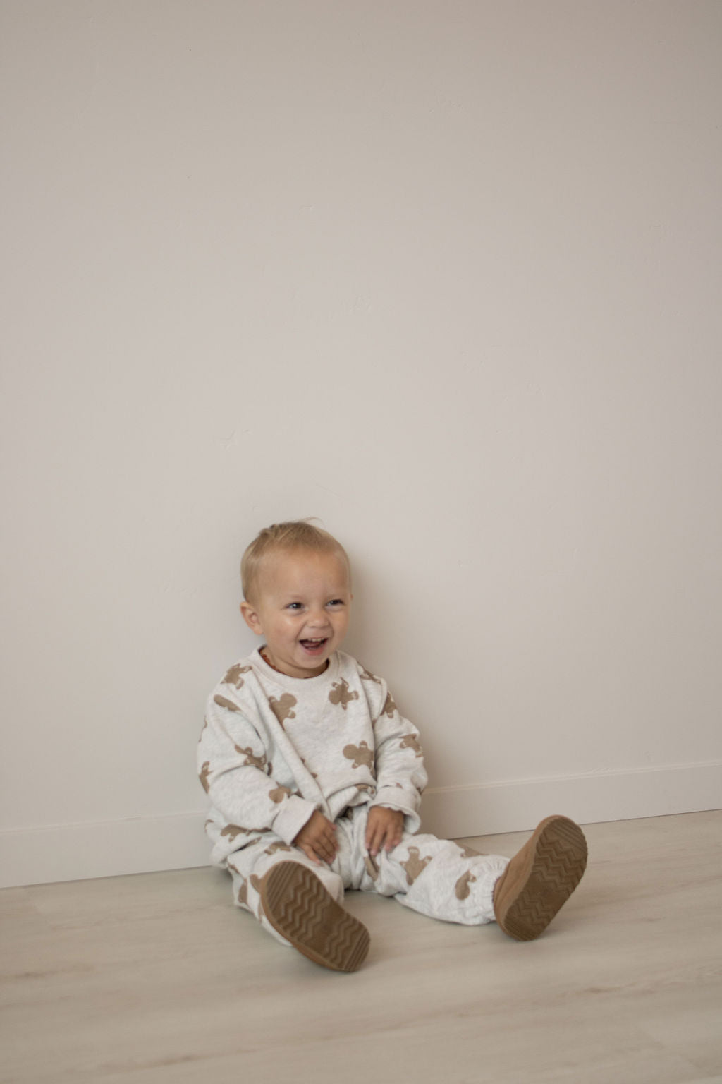 A smiling toddler with short blonde hair sits on a light floor, wearing the forever french baby Sweat Set | Gingerbread 2.0 featuring playful gingerbread-inspired patterns and cozy brown-soled shoes, perfect for everyday adventures.