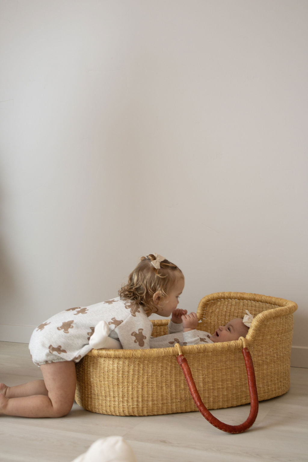 A toddler in a white onesie with brown patterns smiles at a baby wearing the forever french baby Sweatshirt Romper | Gingerbread 2.0, both with light hair and hair bows, indoors against a plain wall.