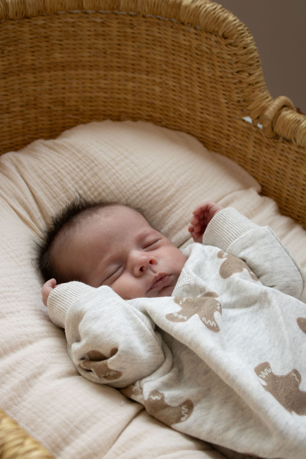 A baby sleeps peacefully on a cream padded blanket in a woven bassinet, wearing the forever french baby Sweatshirt Romper | Gingerbread 2.0 featuring a cozy brown and white bear pattern.