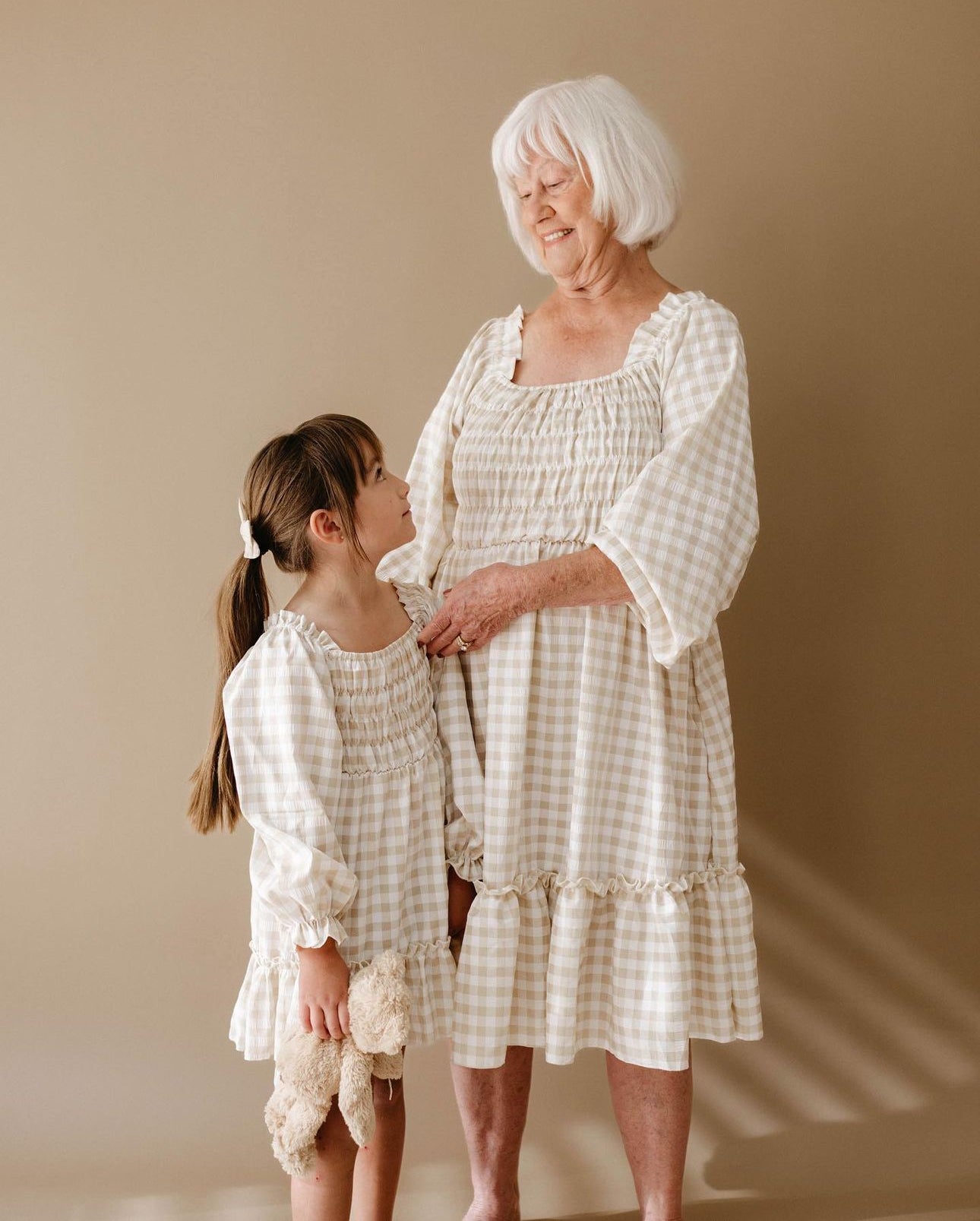 A young girl in a forever french baby Child Dress | Gingham and an elderly woman, both sporting matching outfits, stand side by side. The girl holds onto a stuffed toy while gazing up at the woman, who smiles warmly back at her.