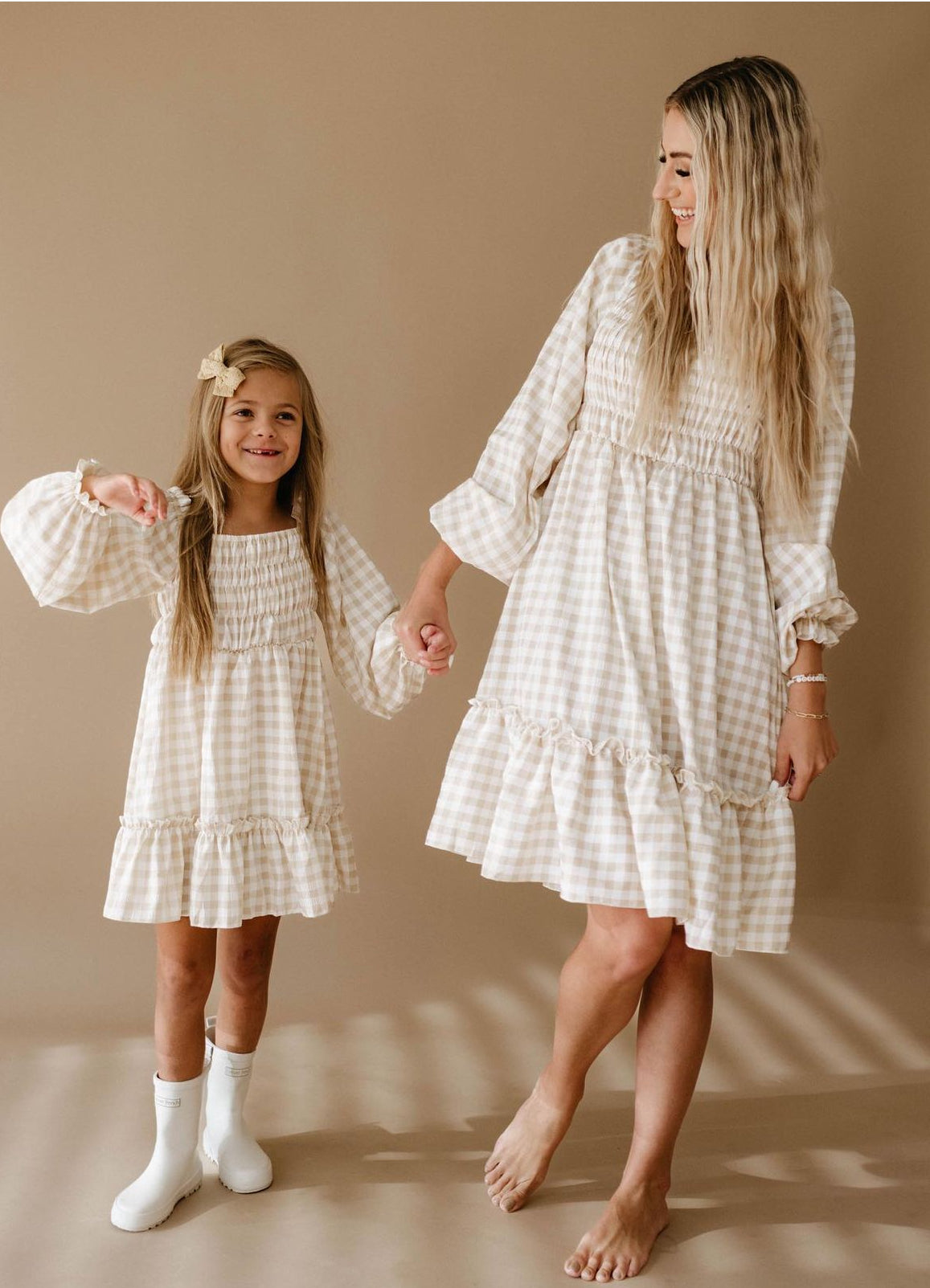 A woman and a young girl, both with long blonde hair, wear matching gingham print dresses from forever french baby's collection. The young girl complements her look with a bow in her hair and white boots. They stand barefoot on a beige background, holding hands and smiling brightly.