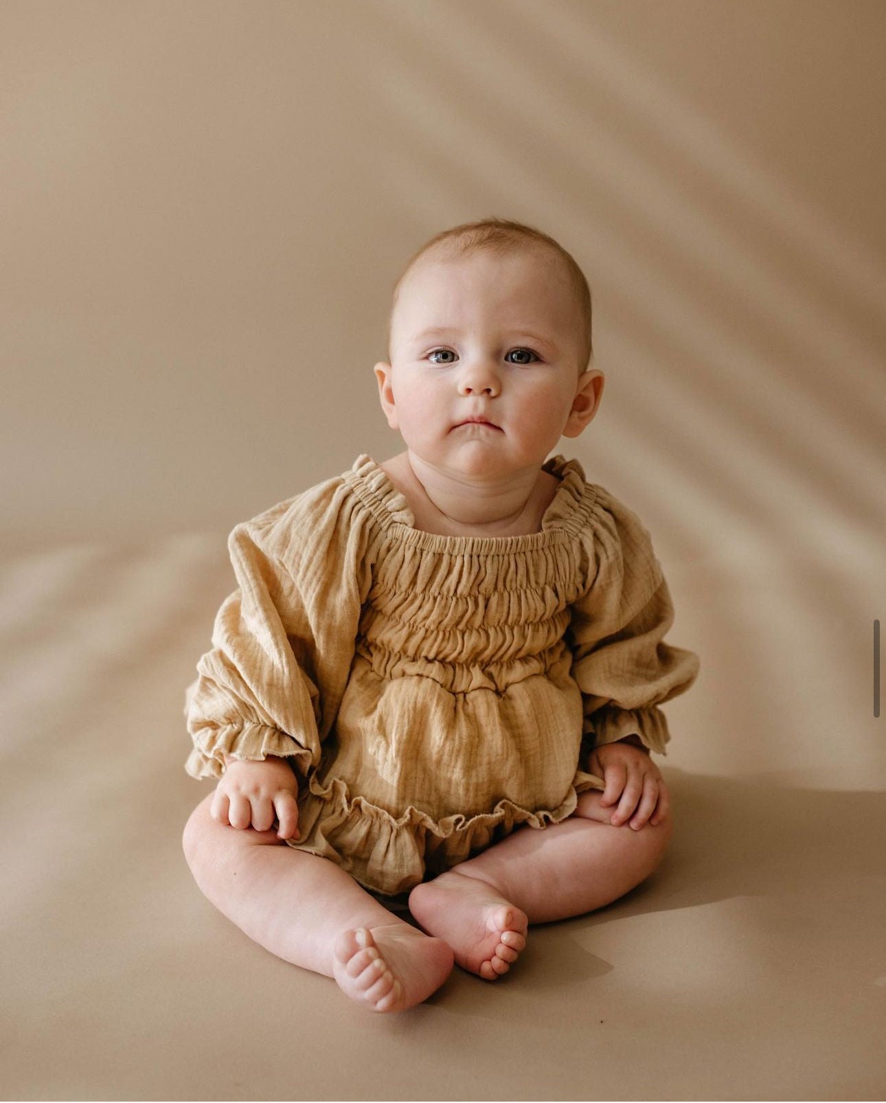 A baby wearing the Ruffle Romper | Golden Hour from forever french baby sits on a matching beige background, looking slightly upward. Their expression is calm and curious, with a golden hue and soft shadows from window blinds gently falling across the scene. This romper is available in sizes ranging from newborn to 24 months.